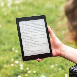A person reads an eBook on a Kobo device while relaxing outdoors in a sunny park.